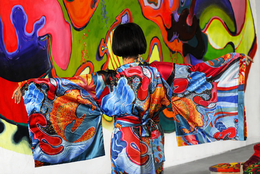 Ornella Ruiz Díaz wearing a hand-painted silk kimono from her phygital series, in front of a large-format mural painting